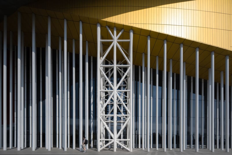 A modern building facade with tall white columns, reflective glass panels, and a yellow curved roof. Two people walk near the base, emphasizing the scale of the structure.