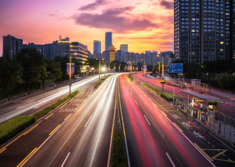 A cityscape at sunset shows tall buildings and a busy, multi-lane road with light trails from fast-moving vehicles, creating streaks of white and red across the scene.