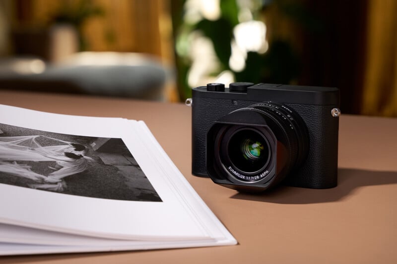 A black digital camera with a large lens sits on a table next to a stack of printed black-and-white photographs. The background is softly blurred with warm tones and green foliage.