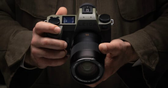 A person in a brown jacket holds a Leica camera with both hands, showing the lens and the top display screen of the camera. The image focuses on the camera and the person's hands.
