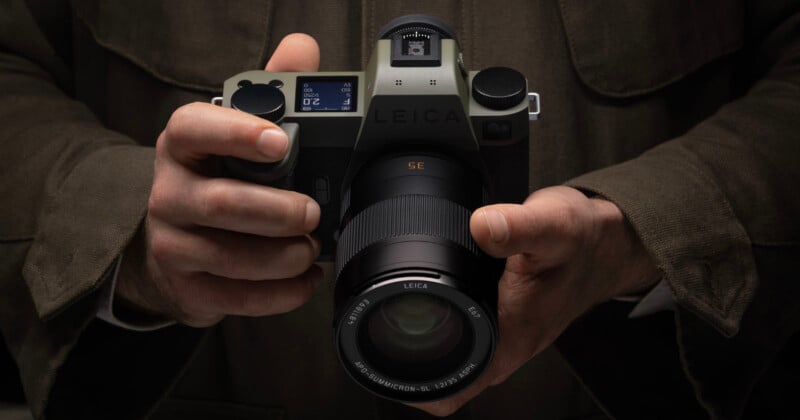 A person in a brown jacket holds a Leica camera with both hands, showing the lens and the top display screen of the camera. The image focuses on the camera and the person’s hands.
