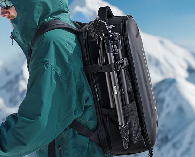 A person in a teal jacket and sunglasses carries a black backpack with a tripod attached, standing in a snowy mountain landscape.