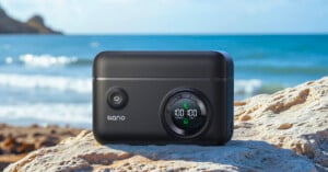A black portable power bank with a circular digital display sits on a sunlit rock near a beach, showing "100" for power and camera, with the sea and blue sky in the background.