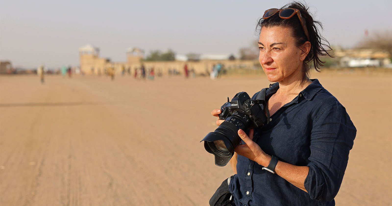 A woman wearing a dark shirt and sunglasses on her head stands on a wide, sandy road holding a camera. The background is blurred with people and buildings visible in the distance.