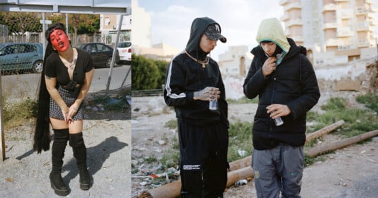 A person with red face paint, black outfit, and boots poses near a fence. Nearby, two people in hooded jackets and streetwear stand on rough ground, drinking water, with buildings in the background.