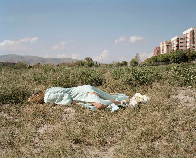A person in a light blue dress lies face down on grass in an open field under a blue sky, with buildings, mountains, and green bushes visible in the distance.