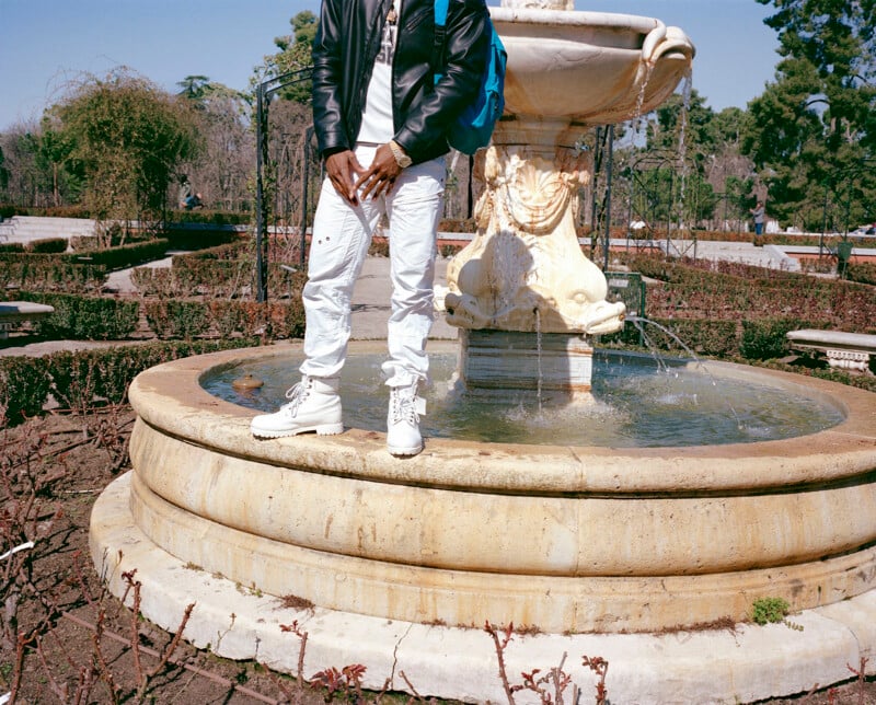 A person dressed in a black jacket, white pants, and white boots stands on the edge of a circular stone fountain in a garden. The person’s head is out of the frame; water flows from the fountain.
