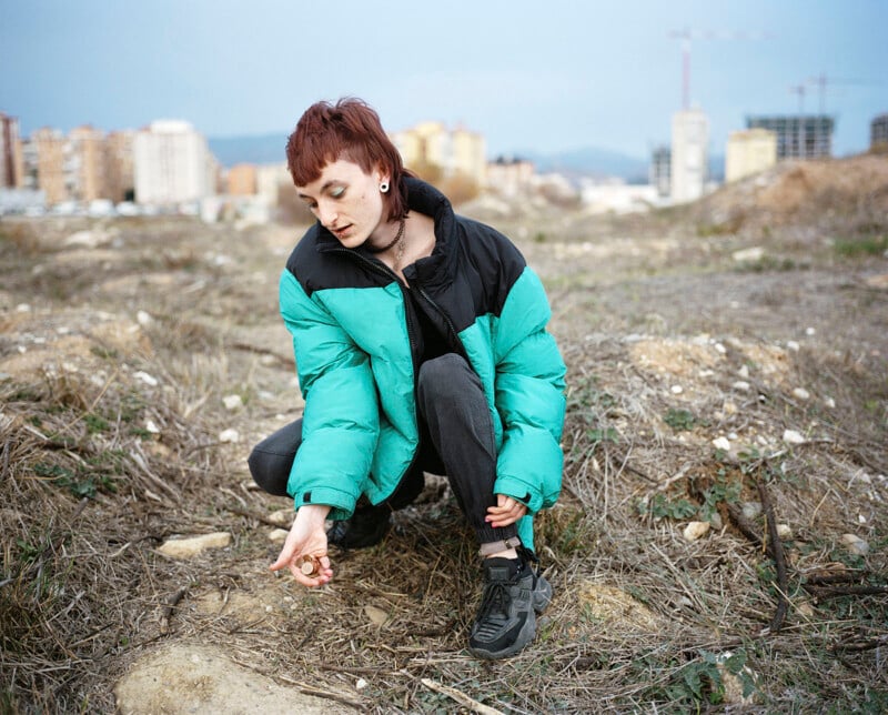 A person with short hair wearing a teal and black puffer jacket squats on a patch of dry grass and dirt, looking down and holding an object, with city buildings and construction cranes in the background.