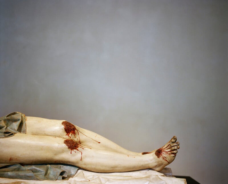 A close-up of a marble sculpture depicting human legs with realistic wounds and blood, lying on a white surface against a plain background.