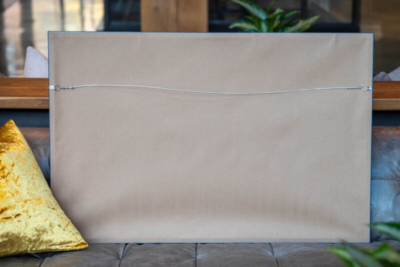A large framed picture turned backward, showing its plain cardboard backing and a hanging wire, rests on a gray leather sofa with a yellow velvet pillow beside it. Some plants and a wooden surface are in the background.