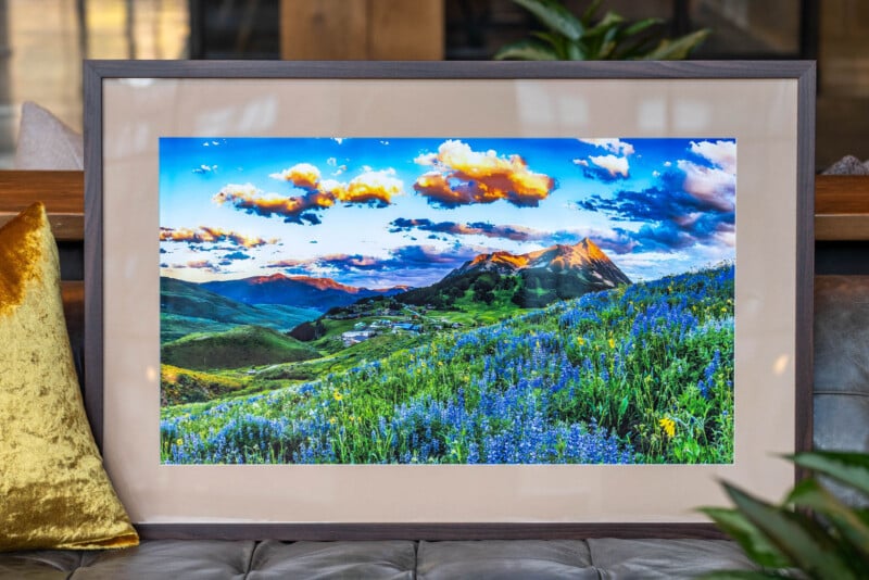 Framed landscape photograph of a mountain scene with a vibrant field of purple wildflowers, rolling green hills, and a colorful sunset sky with clouds, resting on a gray cushioned surface.