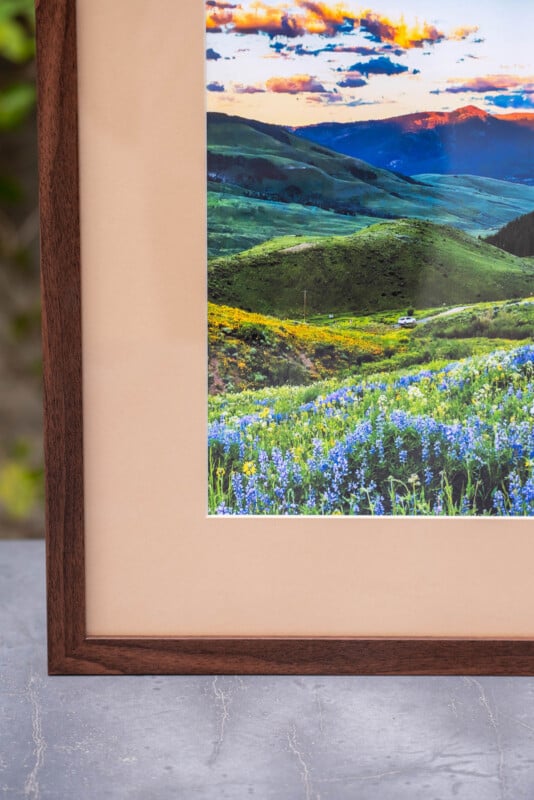 Close-up of a framed landscape photograph showing green hills, wildflowers, and a vibrant sky with clouds at sunset. The wooden frame and a portion of the concrete surface beneath are visible.