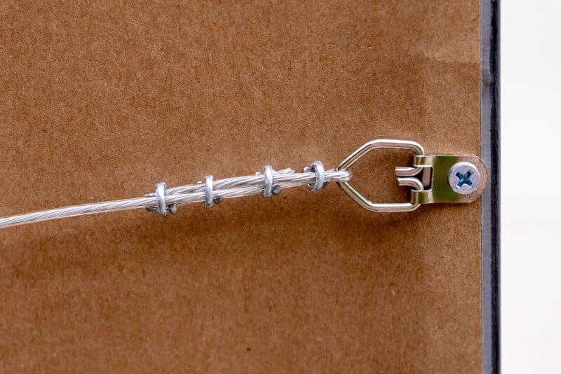 Close-up of a metal picture hanging wire twisted around a hook and secured to the back of a wooden frame with a metal bracket and a screw. The background is brown cardboard.