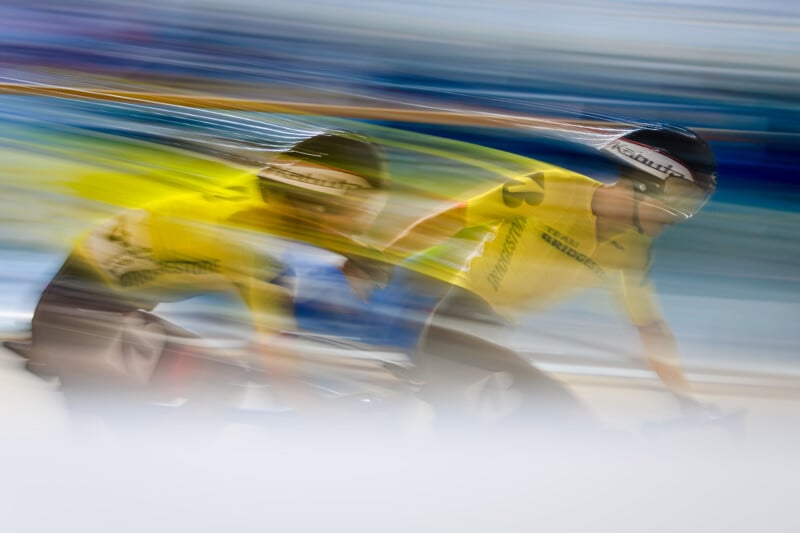 Two cyclists in yellow jerseys and helmets speed around a track, motion blur emphasizing their fast movement and dynamic energy. The background is streaked with blue and yellow, creating a sense of velocity.