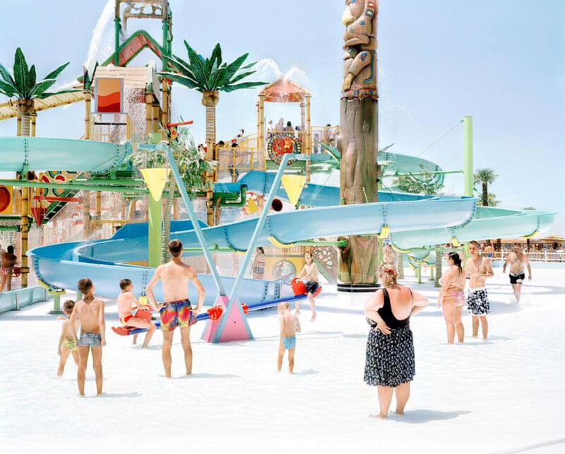 People of various ages stand and play in shallow water at a colorful outdoor water park with slides, palm tree decorations, and fountains under a clear sky.