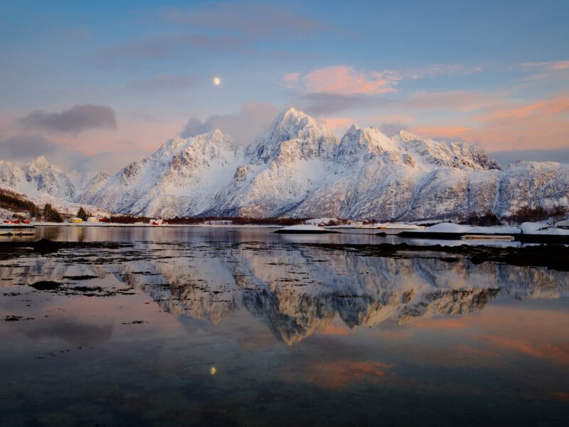 Snow-covered mountains are reflected in a calm, clear lake at sunset. Soft pink clouds streak the sky, and a moon is visible above the peaks. Small houses line the shore in the distance.