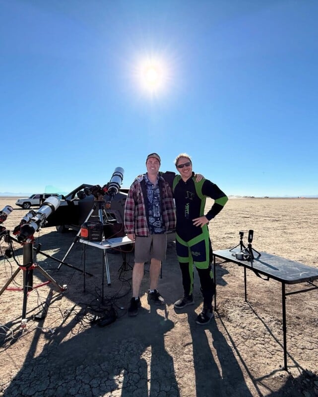 Two people stand smiling in a sunny, barren desert beside telescopes and equipment on tripods. The sun is high in a clear blue sky, and a car is parked in the background.