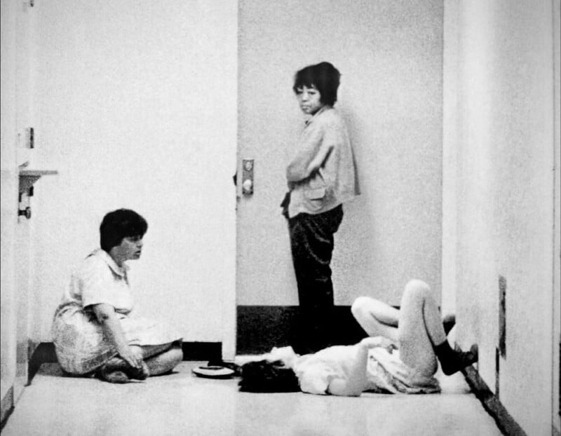 Three women in a bare hallway: one sits on the floor, another stands facing the camera, and the third lies on her back with her legs up against the wall. The scene appears somber and minimalistic.