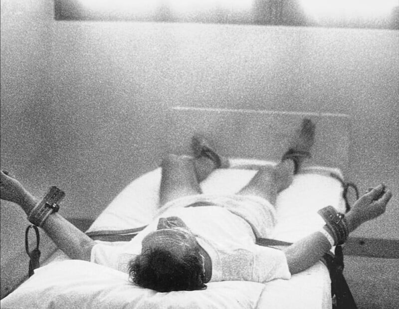 A person lies on a bed in a sparse, bright room, restrained at the ankles and wrists with wide straps. The scene is viewed from above, giving a sense of isolation and vulnerability.