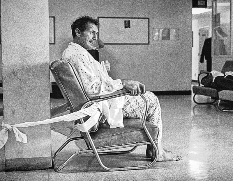 A man in a hospital gown sits barefoot in a chair, with a bed sheet tied from his wrist to the chair’s arm, inside a hospital hallway. He looks sideways and appears to be smiling.
