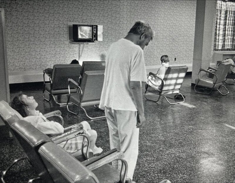 Black-and-white photo of a sparse room with several metal chairs. Three people are present: one sitting with head tilted back, one standing, and one seated near a window. A small TV is on in the background.