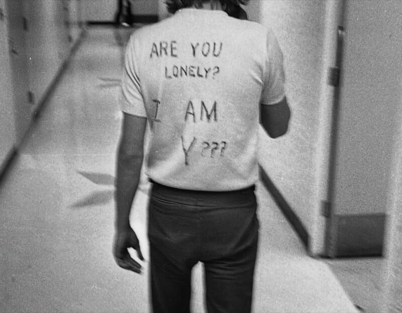 A person walks down a hallway wearing a T-shirt with handwritten text that reads, "ARE YOU LONELY? I AM Y???" The image is black and white and taken from behind.