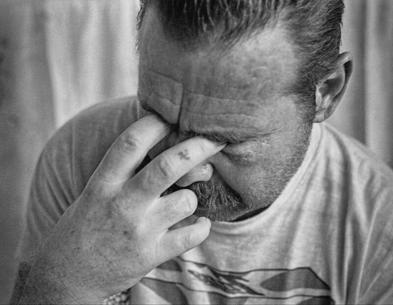 Black and white photo of a man with a mustache, eyes closed, pinching the bridge of his nose with his fingers, appearing stressed or tired. He is wearing a T-shirt and has short hair.