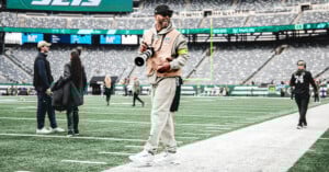 A smiling photographer in casual clothes and a cap stands on the sidelines of a football field holding a camera, with empty stadium seats and a few people in the background.