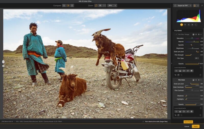 Two people in blue and teal clothing stand on rocky ground next to a motorcycle with a goat sitting on the seat. Another goat lies on the ground, and hills are visible in the background under a partly cloudy sky.