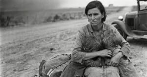 A weary woman in tattered clothes sits on a dirt road, holding two children close as they bury their faces in her lap. An old car is parked nearby in a barren, dusty landscape.