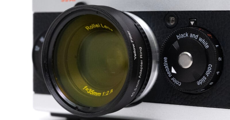 Close-up of a vintage camera lens labeled "Rollei Lens f=35mm 1:2.8" with a yellow filter attached. Next to the lens is a dial labeled "black and white" and "color slide," set on a textured black camera body.