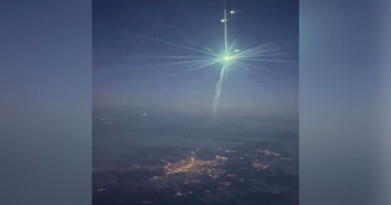 A bright green meteor blazes across the night sky, emitting glowing light with visible streaks, over a dark landscape dotted with city lights far below.