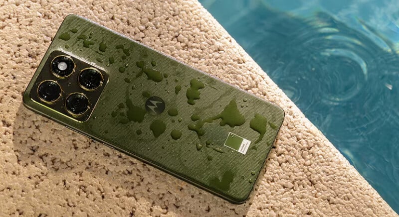 Un teléfono inteligente Motorola verde con gotas de agua en la parte posterior descansa sobre una superficie texturizada junto a una piscina cerca del borde de una piscina.