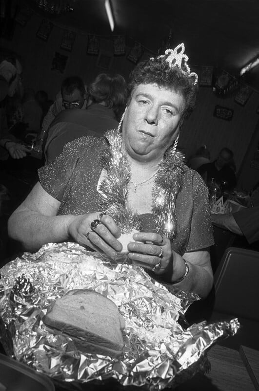 Una persona que lleva una corona, una guirnalda de oropel y una falda brillante está sentada en una habitación llena de gente, sosteniendo un sándwich con pan envuelto en papel de aluminio y luciendo pensativa o pensativa.