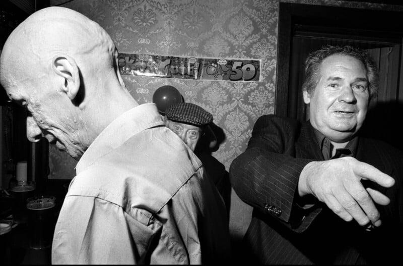 Tres hombres mayores se encontraban en una habitación con poca luz y cubierta con papel tapiz estampado. Una persona está de espaldas a su cara, otra mira a través de la puerta y la otra hace gestos con los brazos. Al fondo se puede ver una pancarta de cumpleaños 