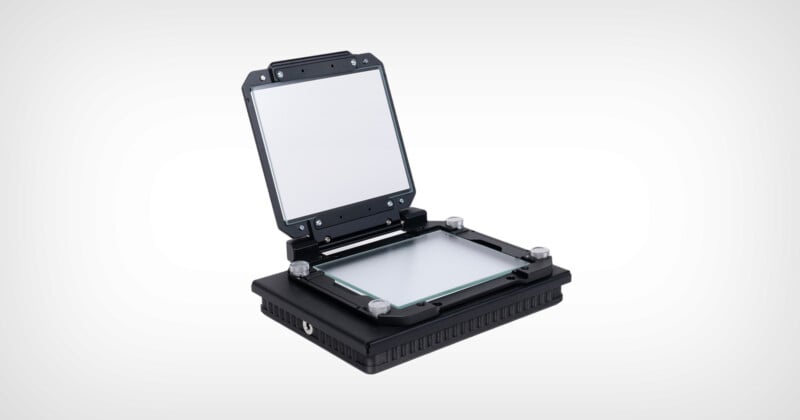 A black, rectangular electronic device with an open hinged lid, revealing a glass surface inside, likely used for holding or aligning materials for processing or inspection, set against a plain white background.