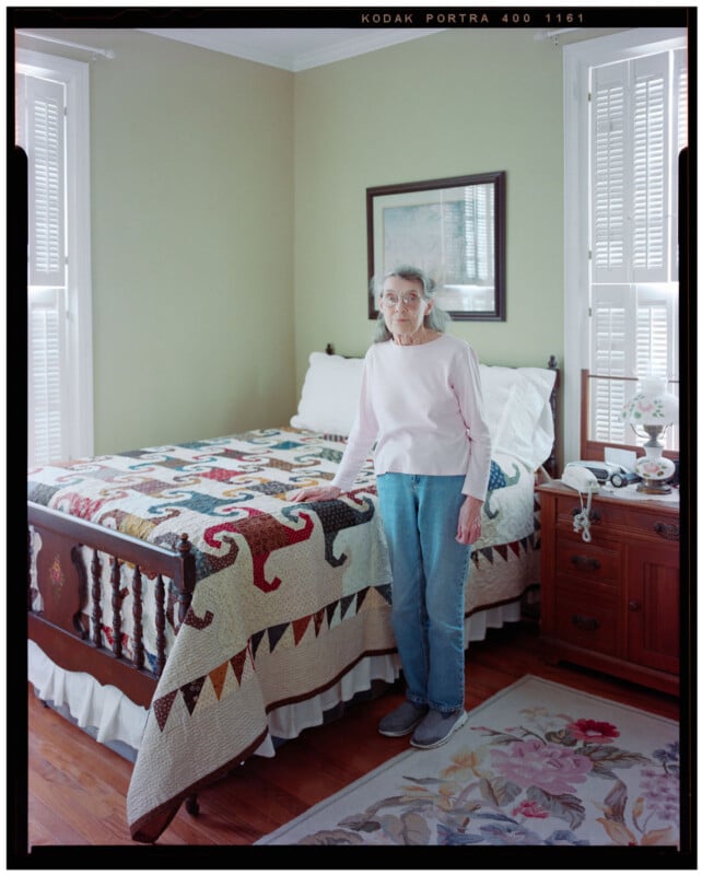 An older woman with gray hair, glasses, a light pink sweater, and blue jeans stands beside a wooden bed with a colorful patchwork quilt in a bright, neatly arranged bedroom.