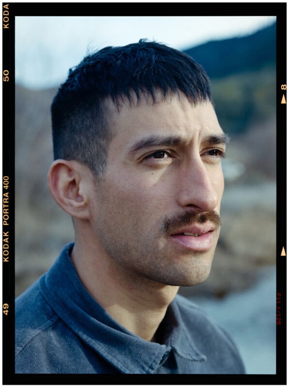 A close-up portrait of a man with short black hair and a mustache, wearing a gray collared shirt, looking slightly to the right outdoors with blurred trees and hills in the background.