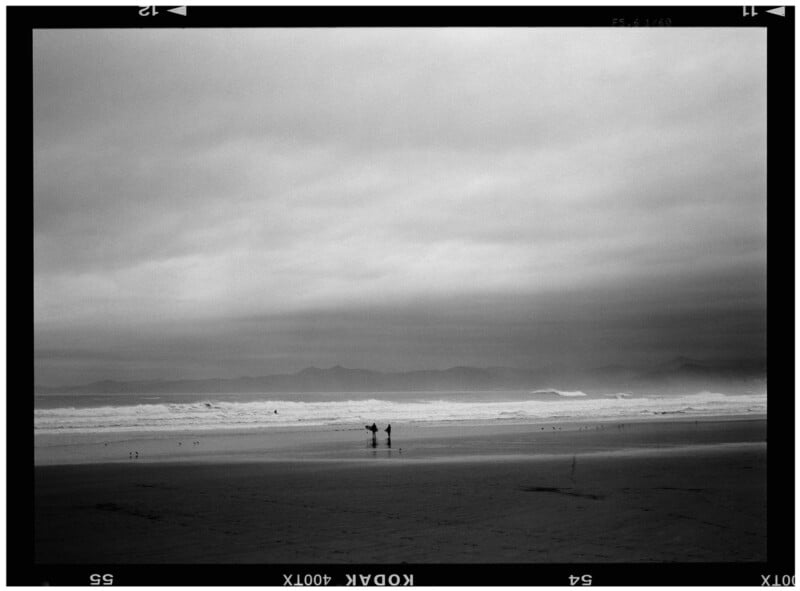 Black and white photo of a beach with two surfers standing near the shore, holding surfboards. Waves crash in the background under a cloudy sky, with mountains faintly visible on the horizon. Film border is visible.