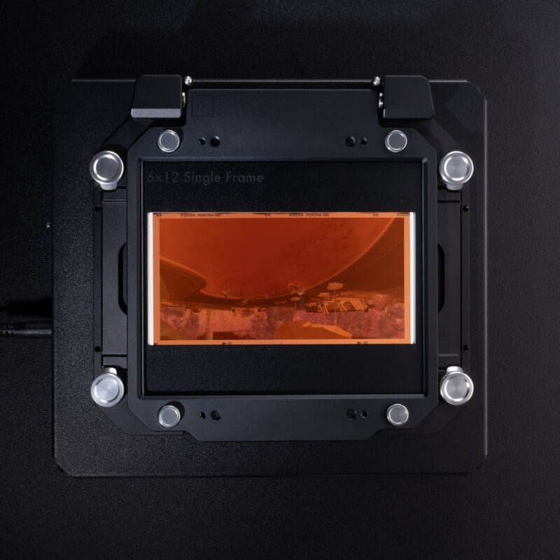 A close-up of a 6x12 film negative placed in a black film holder against a dark background, showing an inverted, orange-tinted image with some buildings and a large structure.
