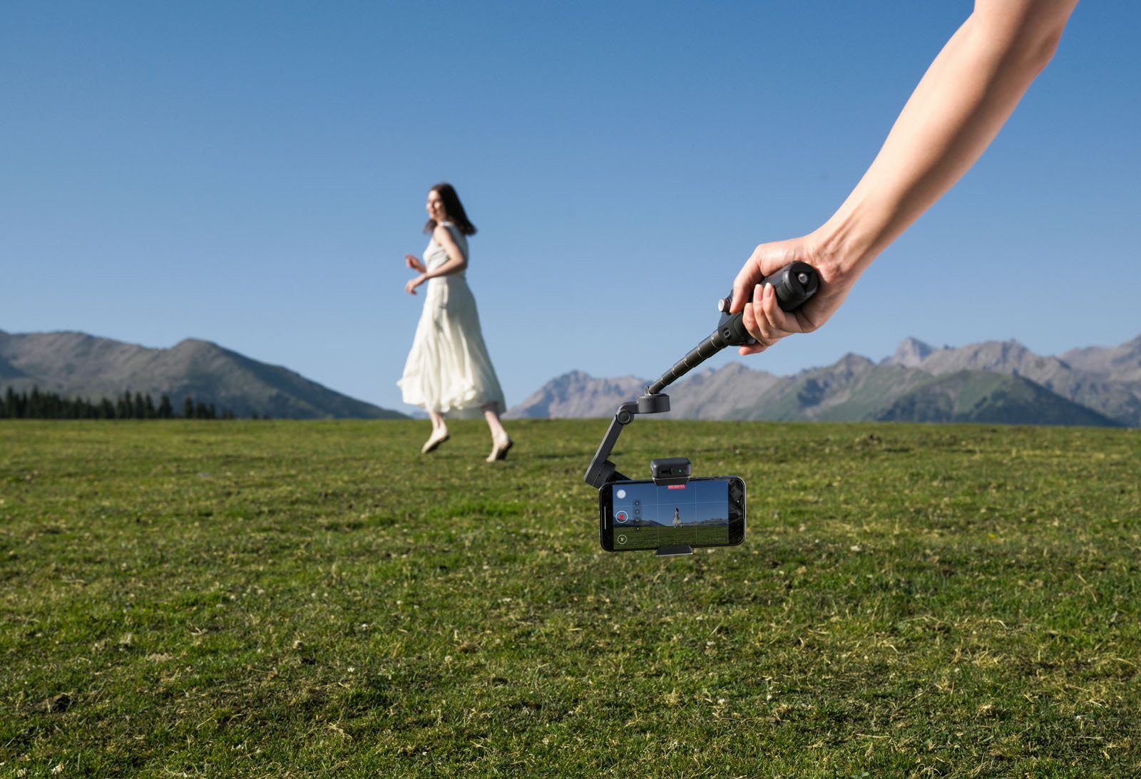 Una persona sostiene un teléfono inteligente montado en un estabilizador y filma a una mujer con un vestido blanco caminando sobre un campo de hierba verde con montañas y un cielo azul claro de fondo.
