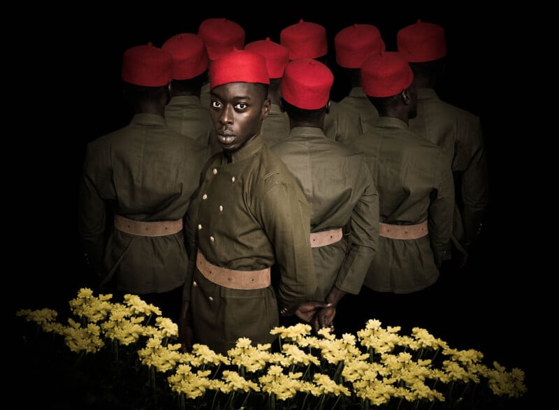 A group of men in olive uniforms and red hats stand in a cluster, most facing away. One man faces forward, gazing at the camera. Bright yellow flowers fill the foreground against a black background.