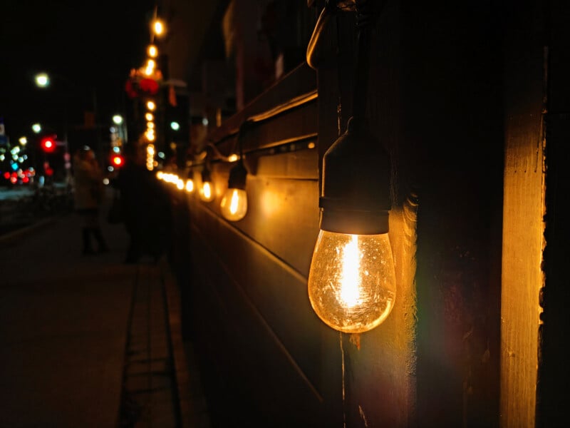 A string of warm glowing light bulbs is hung along a wooden fence at night, illuminating a sidewalk where blurred figures walk in the background.