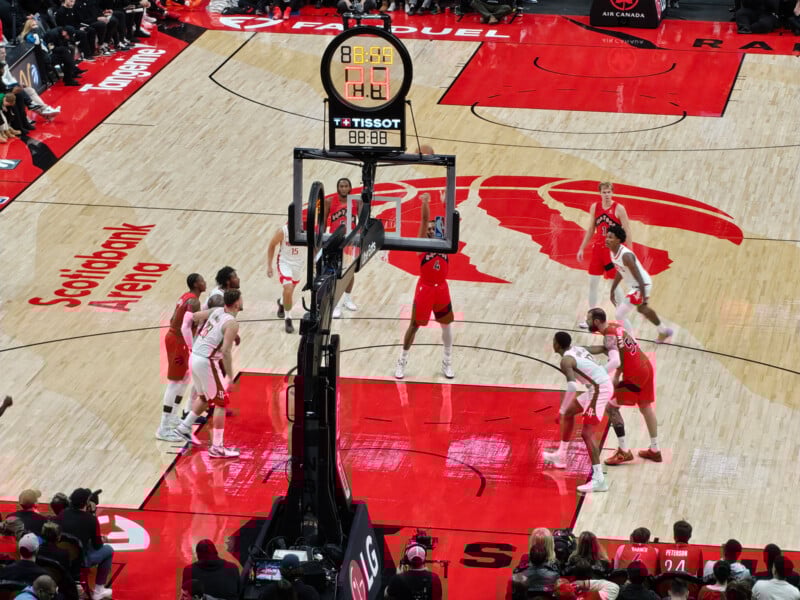 A basketball game at Scotiabank Arena shows players in action near the hoop as a free throw is about to be taken. Spectators are visible in the stands, and the court features a large red Raptors logo.