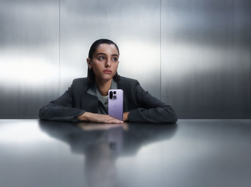 Una mujer con traje gris está sentada en una mesa de metal con un teléfono inteligente lila en posición vertical y una expresión seria sobre un fondo moderno minimalista.
