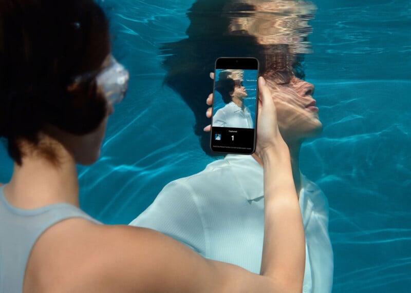 Un hombre sostiene un teléfono inteligente bajo el agua y toma una fotografía de otro hombre que lleva una camisa blanca cerca. La pantalla del teléfono muestra la cara del sujeto bajo el agua, así como su reflejo en la superficie del agua.