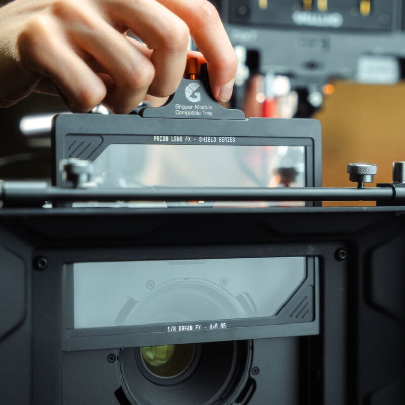 Con una mano se inserta un filtro de lente de prisma rectangular en una caja mate conectada a un equipo de cámara profesional. El texto del filtro dice: "Lente Prisma FX - Serie Escudo" y "1/4 DreamFX - 4x5.65".