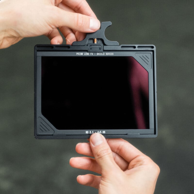 Una persona sostiene un filtro de lente negro rectangular con la etiqueta "Lente prisma serie FX-shield" Manos colocadas sobre un fondo oscuro y borroso.