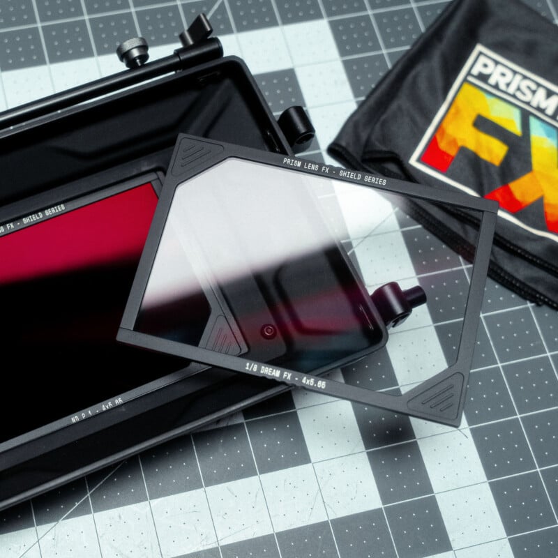 Filtro de lente de cámara rectangular etiquetado "Prisma FX" Colocado en una caja negra con etiqueta en la bolsa. "Prisma FX" junto a ellos. Los artículos están sobre una base de corte gris con un patrón de cuadrícula.