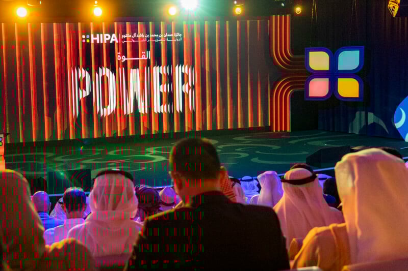 Audience in traditional Middle Eastern attire faces a brightly lit stage with “POWER” and “HIPA” displayed on a large screen, along with Arabic text and colorful geometric designs.